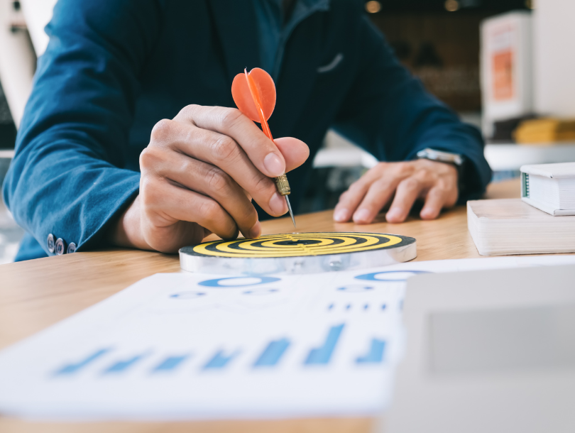 Eine Person in Anzug sitzt an einem Tisch und wirft einen Dartpfeil auf eine Zielscheibe, die auf dem Tisch liegt, neben der sich eine Tabelle mit Statistiken befindet.