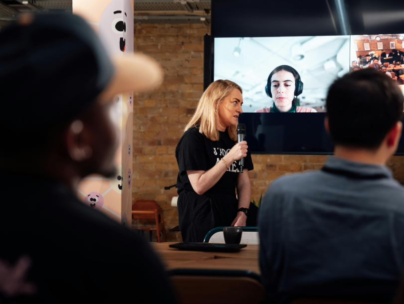Büroumgebung, in der Menschen einer Person zuhören, die mit einem Mikrofon spricht, und auf dem Bildschirm hinter ihnen eine Person mit Kopfhörern ebenfalls zuhört.