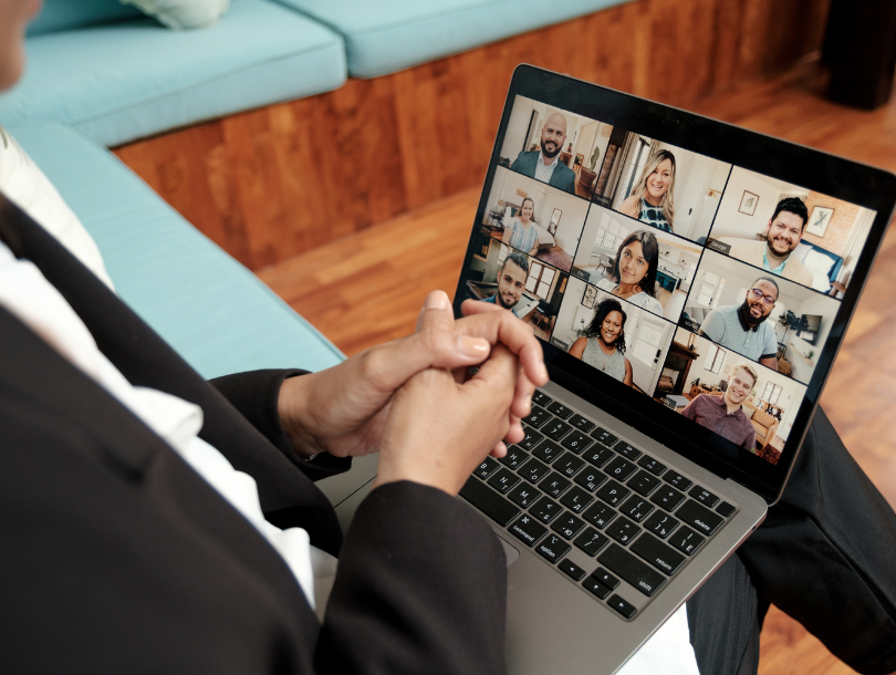 Eine Person mit einem Laptop auf dem Schoß, auf dessen Bildschirm mehrere Personen in kleinen Quadraten zu sehen sind. Eine Art Zoom-Meeting.