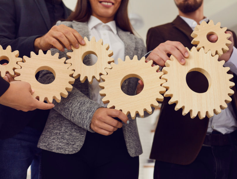4 Personen, die zusammenstehen, halten unterschiedlich große Zahnräder, die ineinander greifen.
