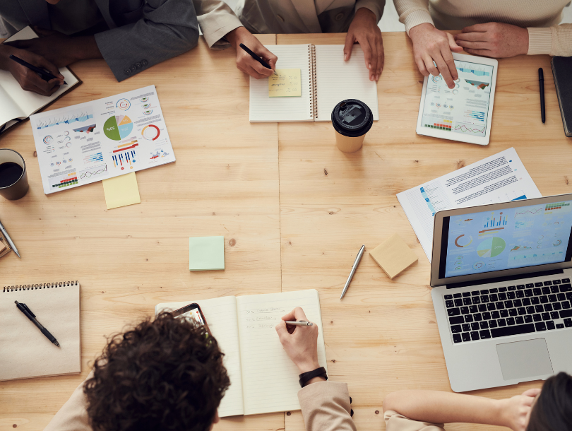 Menschen, die um einen Tisch herum sitzen und eine Besprechung abhalten, bei der man Notizbücher, Laptops und Kaffee sieht.